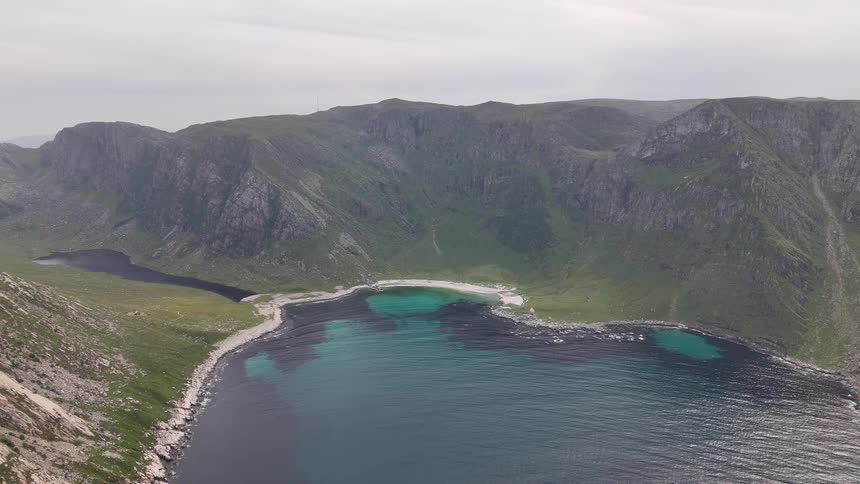 A stunning aerial view shows a secluded cove with a white sand beach and turquoise water, nestled among green mountains in Norway