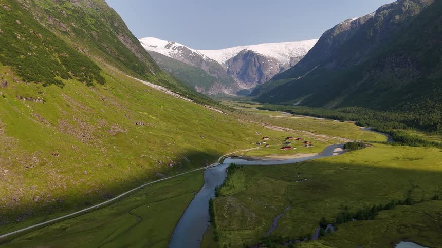 An epic aerial view slowly flies over a lush green valley with a winding river towards a majestic glacier in the distance