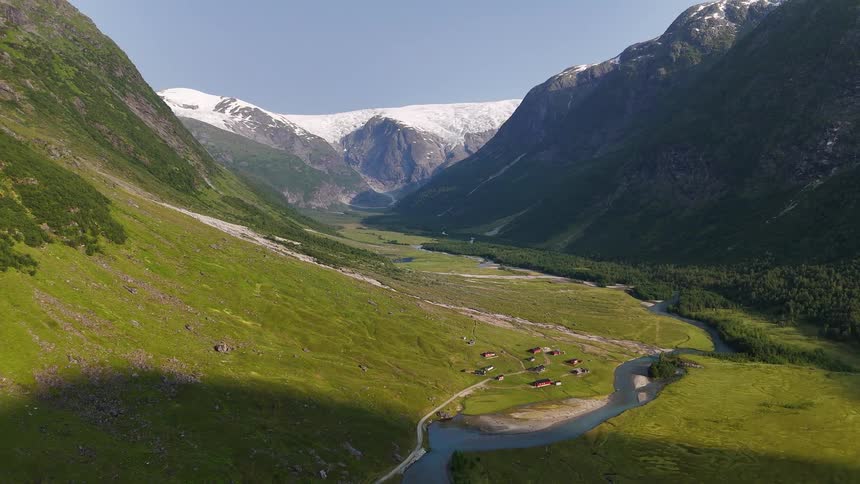 A stunning aerial view of a lush green valley with a river snaking through it, leading to a majestic snow-capped mountain range and a vast glacier