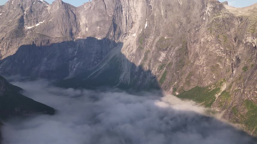 An epic aerial shot tilts up from a cloud-filled valley to reveal the vast, rocky mountains of Trollveggen, Norway