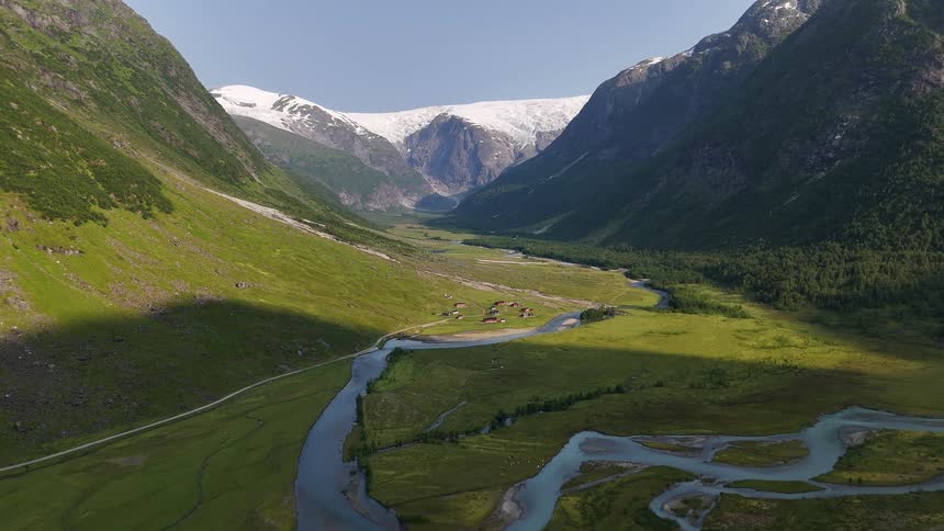 A static aerial view of a beautiful, lush green valley with a winding river leading towards a massive glacier in the mountains