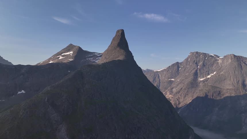 Static aerial shot of a majestic, sharp mountain peak surrounded by a rocky mountain range with patches of snow under a clear blue sky