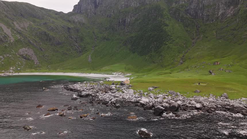 A stationary aerial view showcases the rugged, verdant mountains meeting the rocky coastline and a remote sandy beach with turquoise waters in Lofoten, Norway