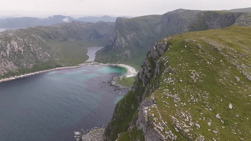 A stunning aerial view from a high cliff reveals a secluded white sand beach and turquoise bay nestled among dramatic green mountains