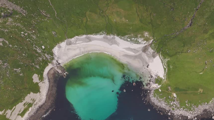 Top-down drone footage slowly rises over a pristine, secluded beach with white sand and clear turquoise water nestled among green, rocky cliffs