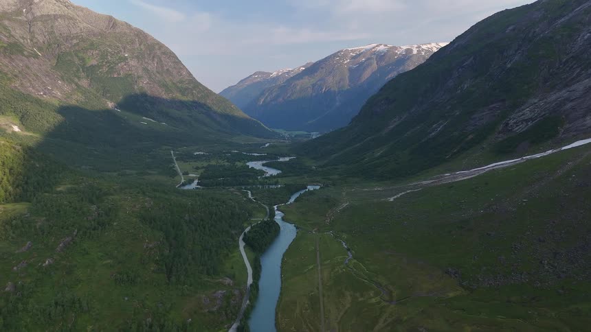 Static aerial shot of a stunning green valley with a winding river flowing between majestic mountains, some with snow-capped peaks