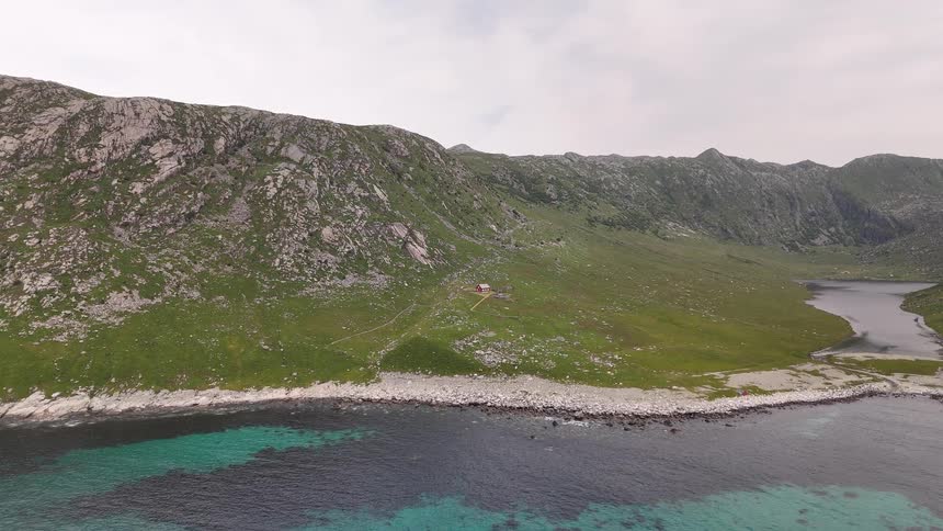 An aerial view slowly moves towards a secluded red cabin on a stunning green coastline with turquoise water and majestic mountains