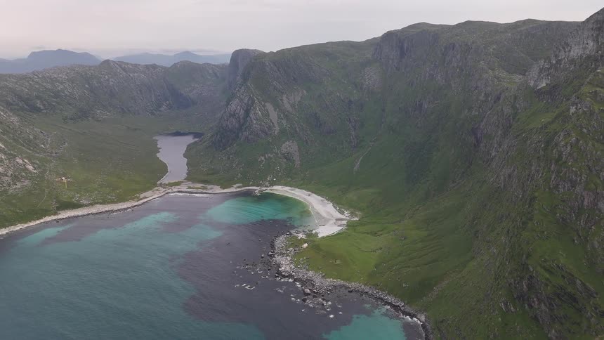 An epic aerial view shows a secluded white sand beach and turquoise bay nestled between lush green mountains and a tranquil lake