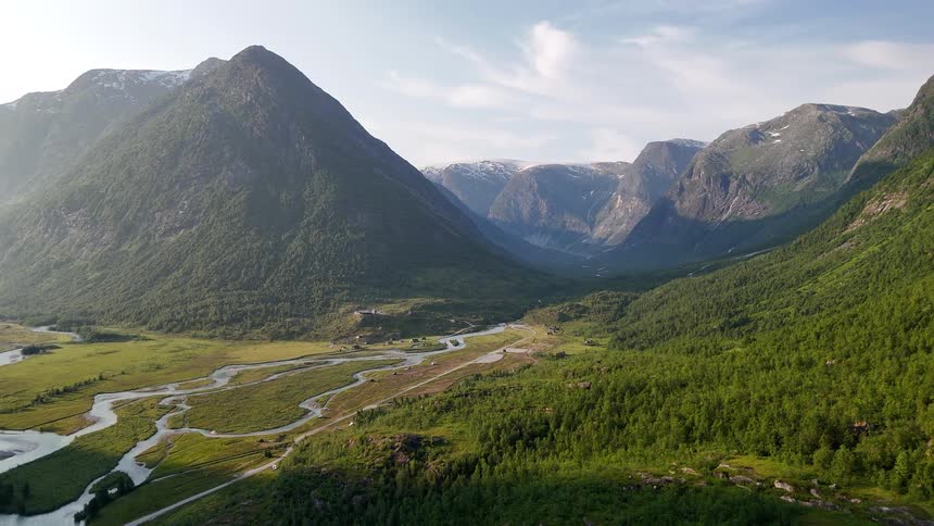 An epic, static aerial shot reveals a stunning mountain valley where a winding river flows through lush green forests and meadows