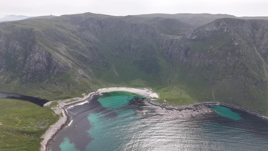 An expansive aerial shot slowly pulls back from a stunningly remote cove with turquoise waters and a white sand beach in the Norwegian mountains