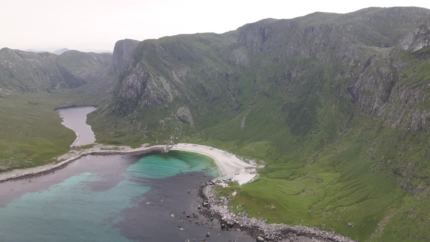 Aerial high-angle view of a secluded white sand beach and turquoise bay surrounded by dramatic green cliffs and a valley lake