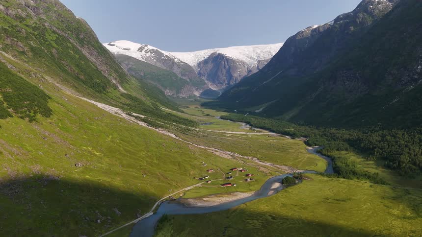 A majestic static aerial view of a verdant Norwegian valley with a winding river, a small village, and a massive snow-covered glacier in the distance