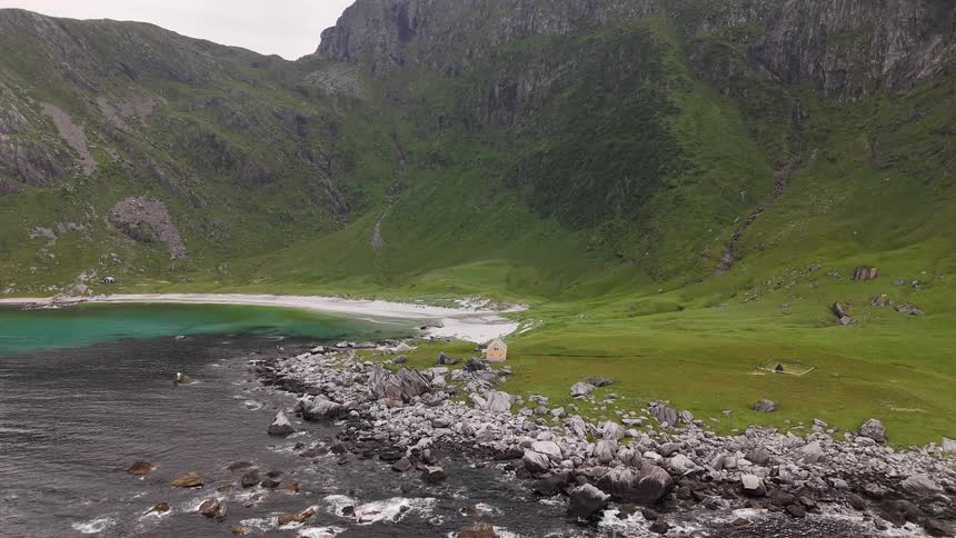 A majestic aerial view reveals a remote Norwegian fjord featuring a secluded white sand beach, turquoise waters, and a lonely cabin surrounded by dramatic green mountains