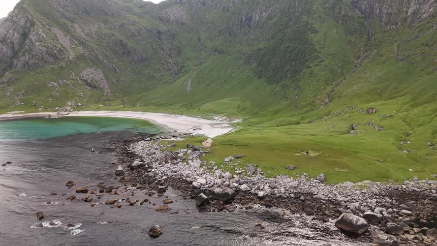A dramatic aerial shot slowly pulls back to reveal a secluded bay with a rocky shore, white sand beach, and vibrant green mountains