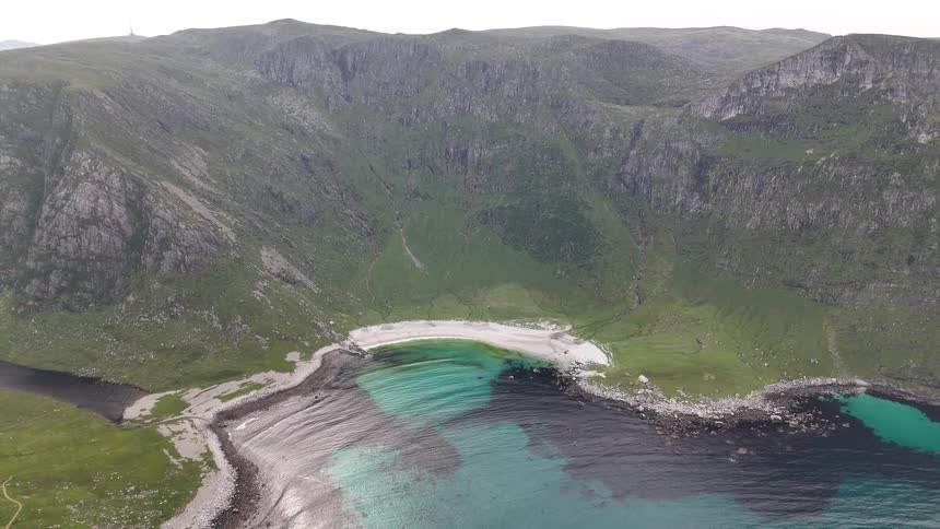 Breathtaking aerial view pulls back from a secluded cove with a white sand beach and crystal-clear turquoise waters surrounded by majestic green mountains