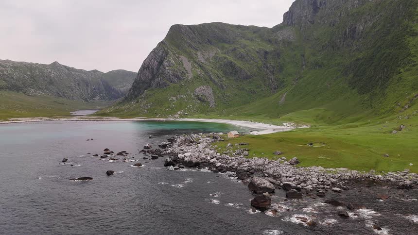 An epic aerial view reveals a remote turquoise water bay and white sand beach tucked away at the base of dramatic green mountains in Norway