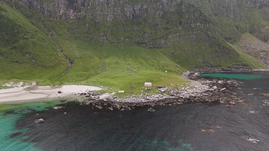 An aerial view reveals a secluded house nestled on a lush green coastline next to a sandy beach and turquoise waters at the base of a majestic mountain