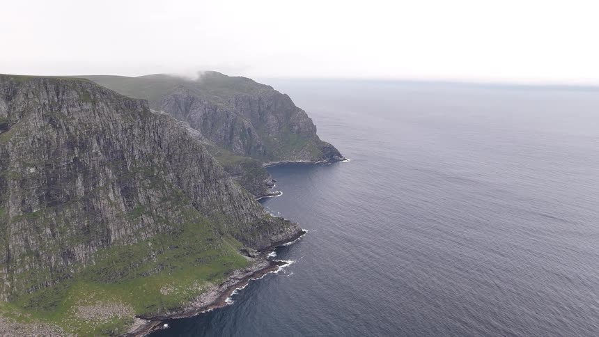 A static aerial view shows the majestic, rugged cliffs of a green coastline meeting the vast, calm ocean on a cloudy day