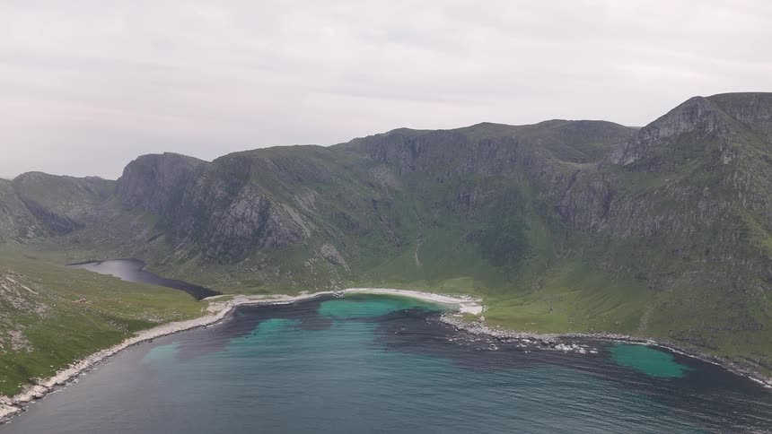 A stunning aerial view shows a secluded bay with turquoise waters, a white sand beach, and a hidden lake nestled among towering green mountains