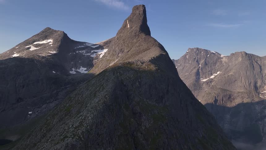 A slow aerial shot pulls back to reveal the iconic and rugged Romsdalshorn mountain peak in Norway under a clear blue sky