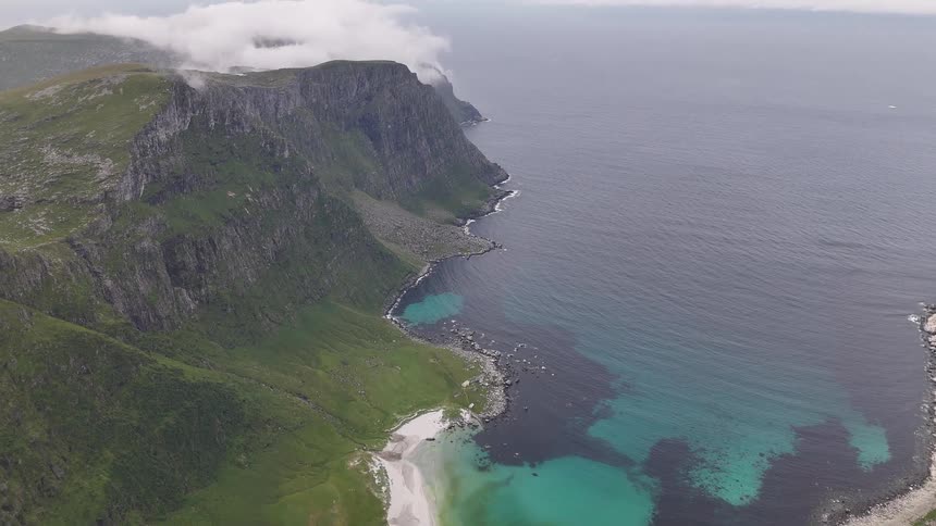 An epic aerial view tilts up from a secluded white sand beach and turquoise cove to reveal the vast ocean and majestic, cloud-covered mountains