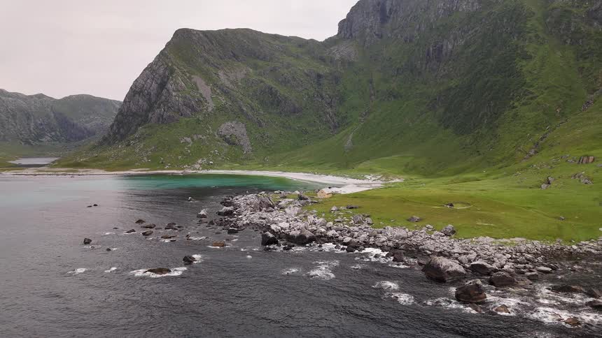 An aerial view of a remote and scenic Norwegian coastline with turquoise waters, a white sand beach, and lush green mountains
