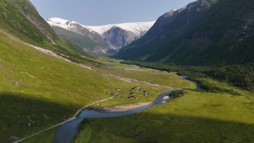 An epic aerial shot pushes forward over a lush green valley with a winding river and a small village, revealing a massive glacier in the background