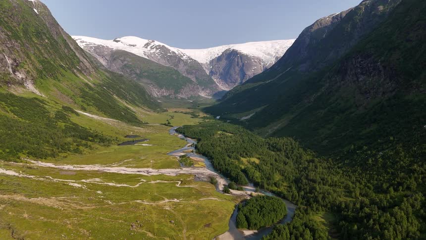 Aerial view flying over a vast green valley with a winding river leading to snow-covered mountains and a glacier in Norway