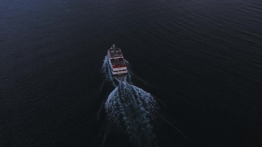 An aerial view shows a tour boat cruising through the dark, calm waters at twilight, leaving a white wake behind it