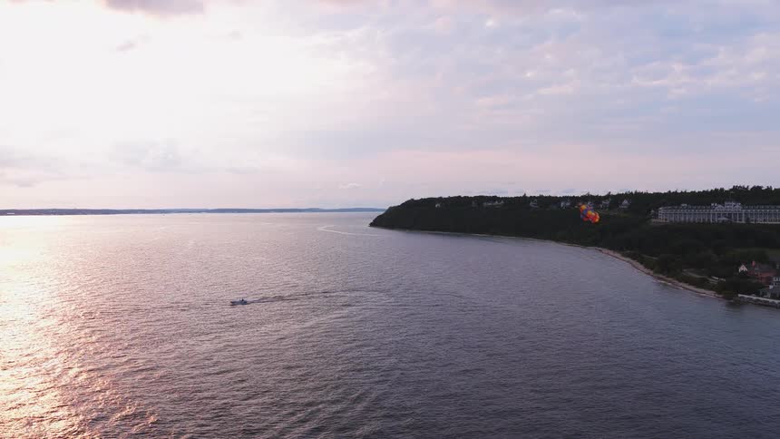 An aerial view captures a boat parasailing over the water during a beautiful sunset along a scenic, tree-lined coastline