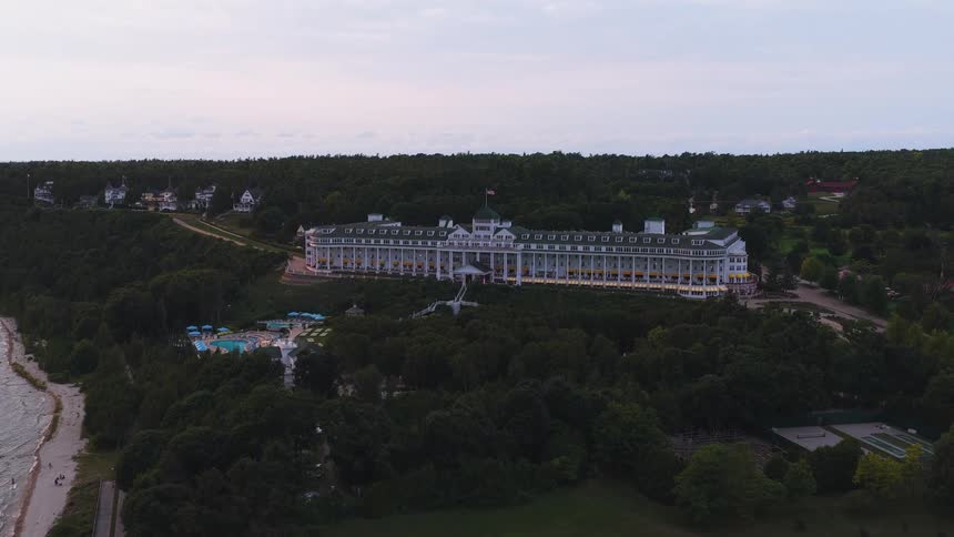 An enchanting aerial push-in shot of the historic Grand Hotel, perched on a forested bluff above the Great Lakes coastline during twilight