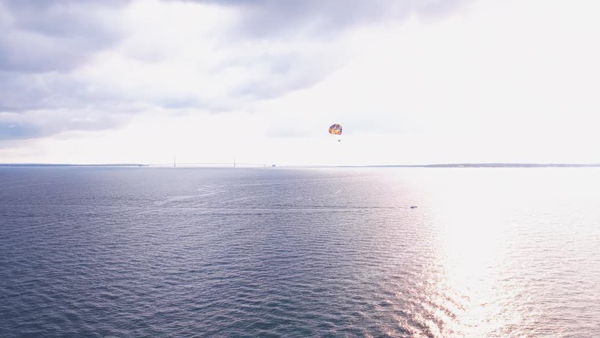 An aerial view captures a serene parasailing adventure over a vast lake with a distant suspension bridge silhouetted against a bright, cloudy sky