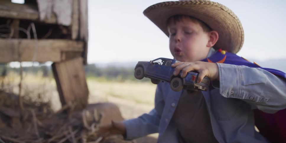 A young boy in a cowboy hat and a superhero cape plays with his toy truck in the dirt in slow motion