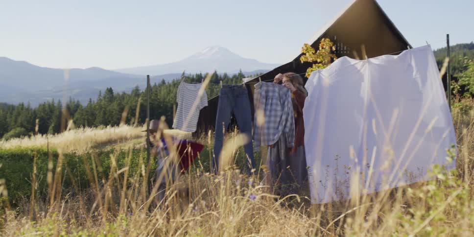 A mother hangs laundry on a clothesline in a sun-drenched rural field while her young son plays in his cape, with a mountain in the distance