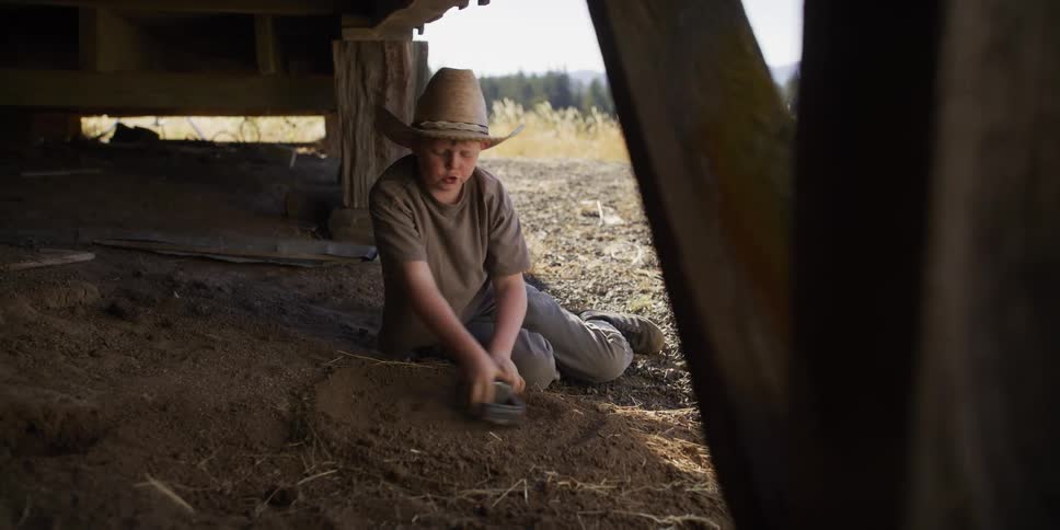 A young farm boy wearing a cowboy hat sits in the dirt under a barn and plays with his blue toy truck