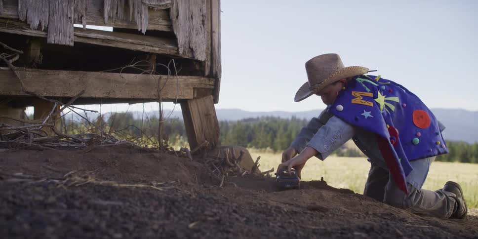 A young boy in a cowboy hat and a superhero cape runs in slow motion to play with his toy truck in the dirt by an old barn
