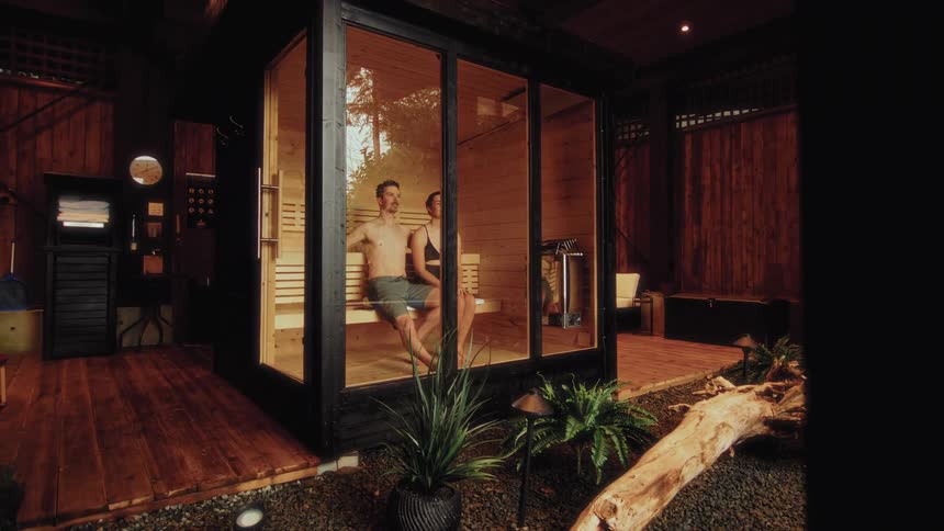 A couple relaxes together inside a modern glass sauna in a tranquil, beautifully designed spa-like setting