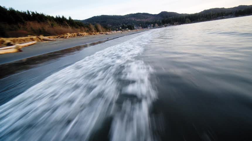 A dynamic low-angle aerial shot flies swiftly along the shoreline, following a wave washing onto the sand