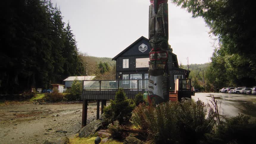A tranquil scene of a coastal lodge with a traditional Indigenous totem pole during a light snowfall in a remote, forested location