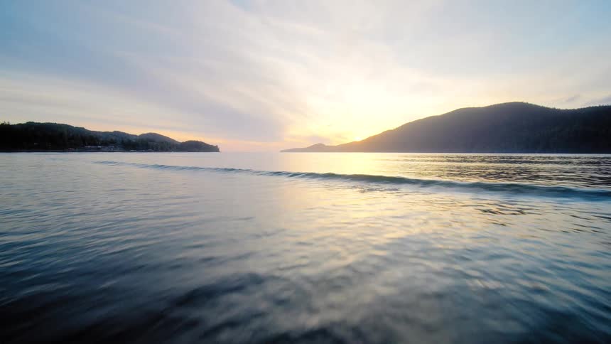 A low-flying aerial shot glides from a sandy beach over gentle ocean waves towards a beautiful golden sunset behind distant mountains