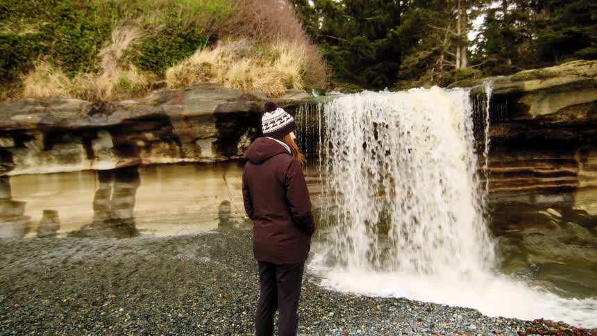 A person in a winter hat stands on a rocky beach, mesmerized by a waterfall cascading over a coastal cliff in this cinemagraph