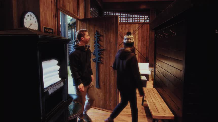 A couple in winter attire enters the warm and inviting wooden anteroom of a sauna at a luxury spa