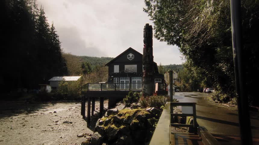 A slow forward dolly shot unveils a First Nations-style building with a totem pole on a rocky shore surrounded by a dense forest