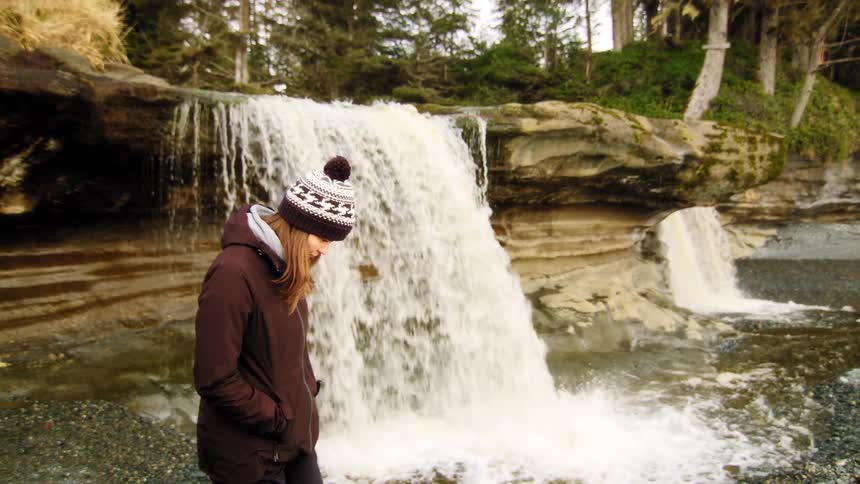 A contemplative woman wearing a beanie and jacket stands beside a flowing waterfall in a natural coastal forest