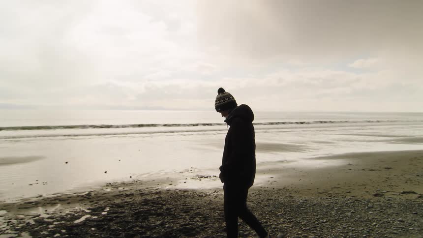 A lone person in winter clothing takes a thoughtful, slow-motion walk along a desolate beach on a cloudy day