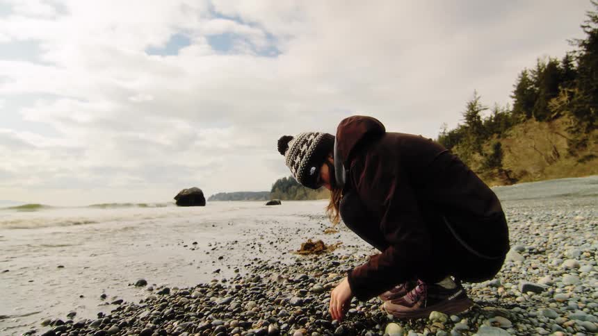 A woman in a warm hat and jacket crouches down to explore the rocky shoreline of a cool beach as waves gently lap the shore
