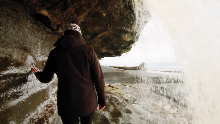 A woman in a winter coat and hat stands in a cave behind a waterfall, looking out at the rocky beach in slow motion