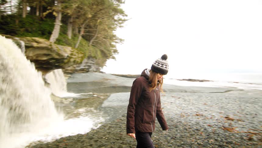 A woman in a winter hat and jacket walks along a rocky beach past a beautiful waterfall flowing into the ocean