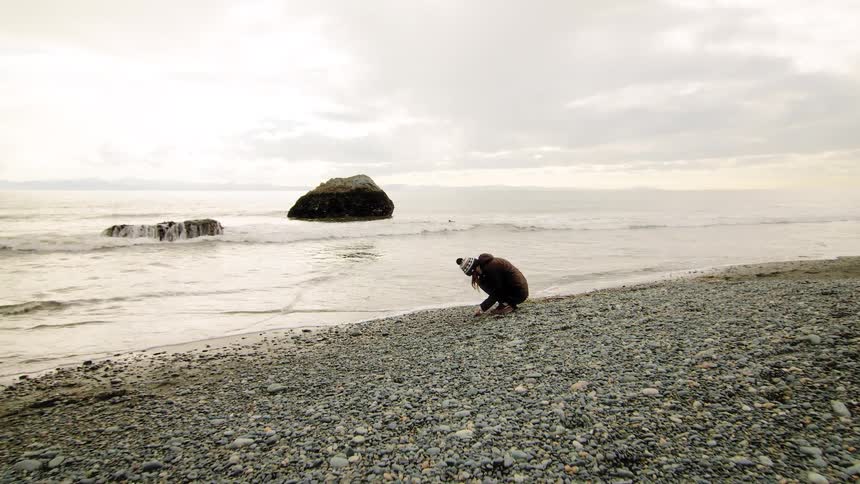 A wide shot shows a photographer in a beanie capturing the serene coastal landscape on a rocky beach