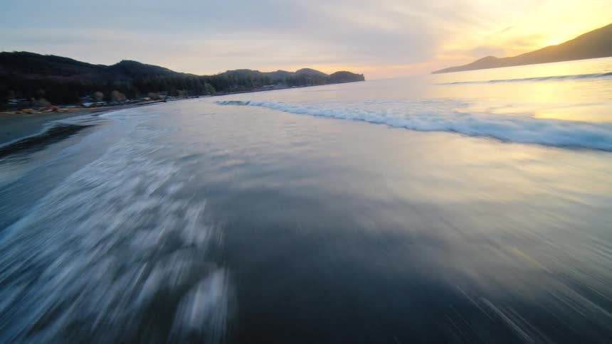 A dynamic aerial shot races along the shoreline with a breaking wave before soaring out over the ocean towards a tranquil sunset